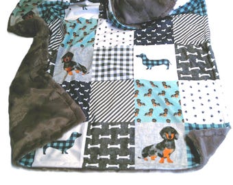 May include: A patchwork blanket featuring a variety of square fabric panels with dachshund dog patterns, black and white stripes, and bone motifs. The blanket has a soft, dark gray plush backing and a hood, with blue, black, and white color tones.