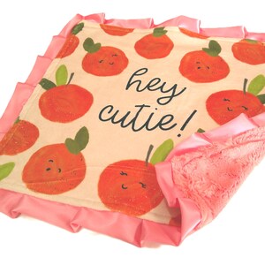 May include: A square baby security blanket featuring a cream-colored top with a repeating pattern of smiling orange citrus fruits and green leaves. The text "hey cutie!" is printed in black. The blanket has a pink satin ruffle trim and a textured pink backing.
