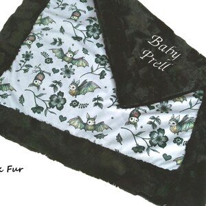 May include: A soft blanket featuring a white fabric center with a repeating pattern of green bats, dark flowers, and small hearts. The blanket is bordered by a thick, plush black fur trim. The text Baby Prell is embroidered in white on the black fur.