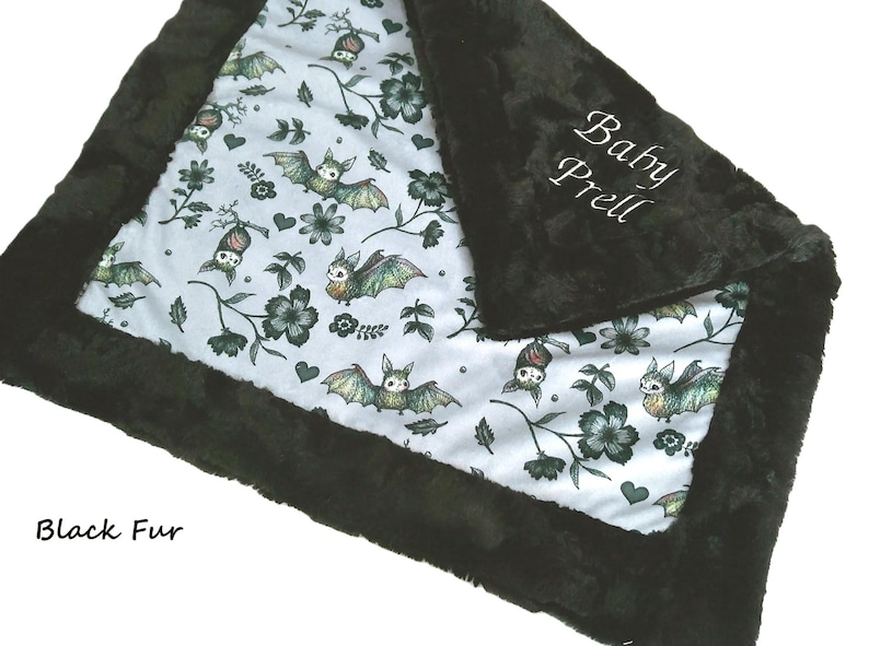 May include: A soft blanket featuring a white fabric center with a repeating pattern of green bats, dark flowers, and small hearts. The blanket is bordered by a thick, plush black fur trim. The text Baby Prell is embroidered in white on the black fur.