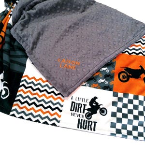 May include: A gray and multi-colored patchwork blanket featuring motorcycle graphics and the text "A Little Dirt Never Hurt." The blanket has a gray, dimpled top with the embroidered text "CAISON LANE."