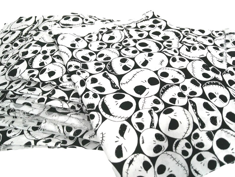 May include: White and black fabric featuring a repeating pattern of Jack Skellington's face from The Nightmare Before Christmas. The fabric is folded, showing multiple layers of the printed design. The design is a popular theme for Halloween and spooky decor.