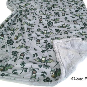 May include: A light gray blanket featuring a repeating pattern of green and brown bats, dark flowers, and small hearts. The blanket is bordered by a textured silver faux fur trim. The text Silver Fur is printed in the bottom right corner.