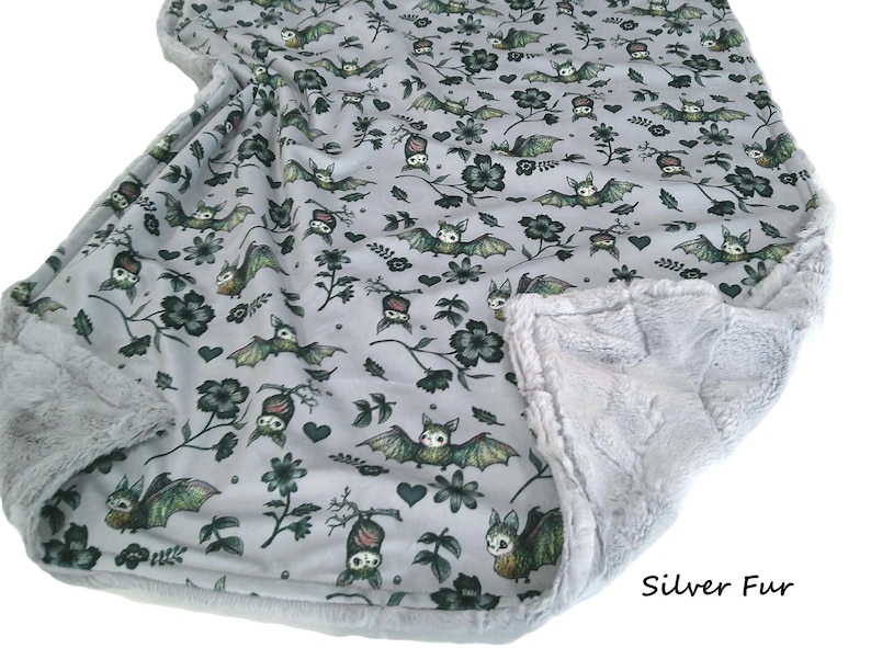 May include: A light gray blanket featuring a repeating pattern of green and brown bats, dark flowers, and small hearts. The blanket is bordered by a textured silver faux fur trim. The text Silver Fur is printed in the bottom right corner.