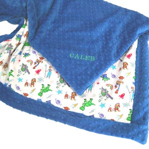 May include: A blue dimpled minky blanket featuring a white underside with a colorful cartoon character print including space themes and toys. The name CALEB is embroidered in green thread on the blue side of the folded fabric corner.
