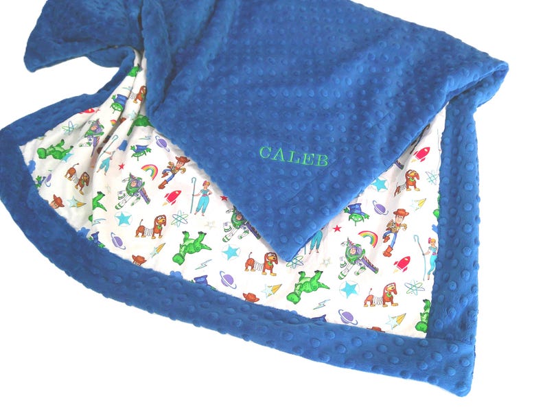 May include: A blue dimpled minky blanket featuring a white underside with a colorful cartoon character print including space themes and toys. The name CALEB is embroidered in green thread on the blue side of the folded fabric corner.
