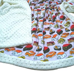 May include: A soft blanket featuring a light pink background with a repeating pattern of cats shaped like sushi rolls. The blanket has a textured cream-colored minky dot fabric border that frames the central sushi cat print on the front side.
