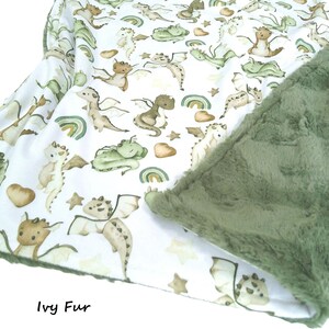 May include: A baby blanket with a white background featuring a pattern of watercolor dragons in shades of green, brown, and tan. The blanket has a soft, green, plush backing. The text "Ivy Fur" is visible.