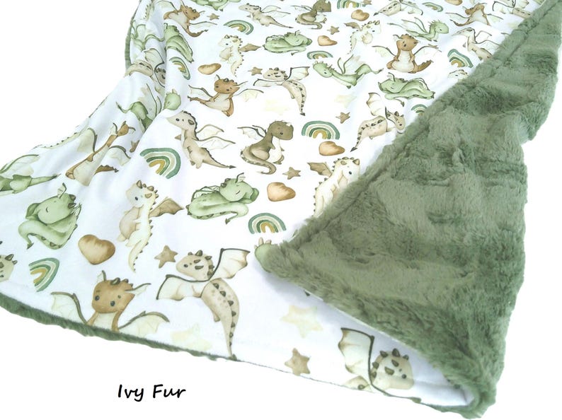 May include: A baby blanket with a white background featuring a pattern of watercolor dragons in shades of green, brown, and tan. The blanket has a soft, green, plush backing. The text "Ivy Fur" is visible.