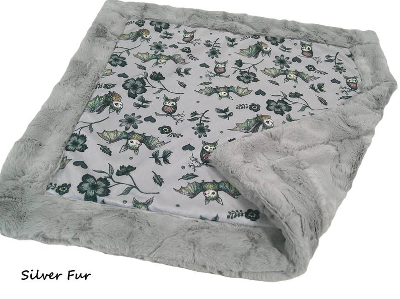 May include: A rectangular blanket featuring a light gray center panel with a repeating pattern of bats, owls, and dark green flowers. The blanket is bordered by a thick, textured silver faux fur trim. The text Silver Fur is visible.