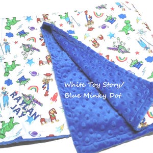 May include: A baby blanket featuring a white side with a colorful Toy Story character print and the name RAYLAN JAY embroidered in blue, and a solid blue minky dot textured side. The text White Toy Story/Blue Minky Dot is visible.