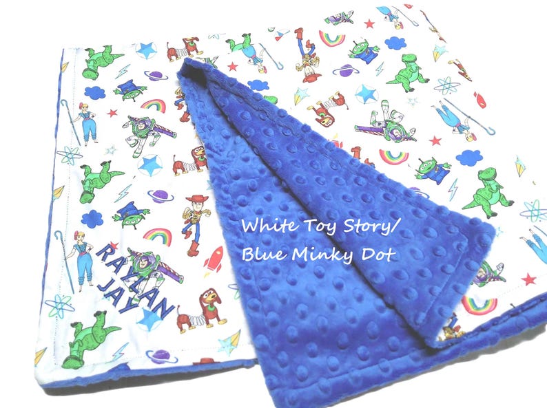 May include: A baby blanket featuring a white side with a colorful Toy Story character print and the name RAYLAN JAY embroidered in blue, and a solid blue minky dot textured side. The text White Toy Story/Blue Minky Dot is visible.