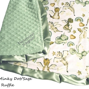 May include: Sage green baby blanket with a dragon pattern. The blanket features a soft, textured minky dot fabric on one side and a dragon print on the other. The edges are trimmed with a sage satin ruffle. The name "Desmond Vagnier" is embroidered on the minky side.