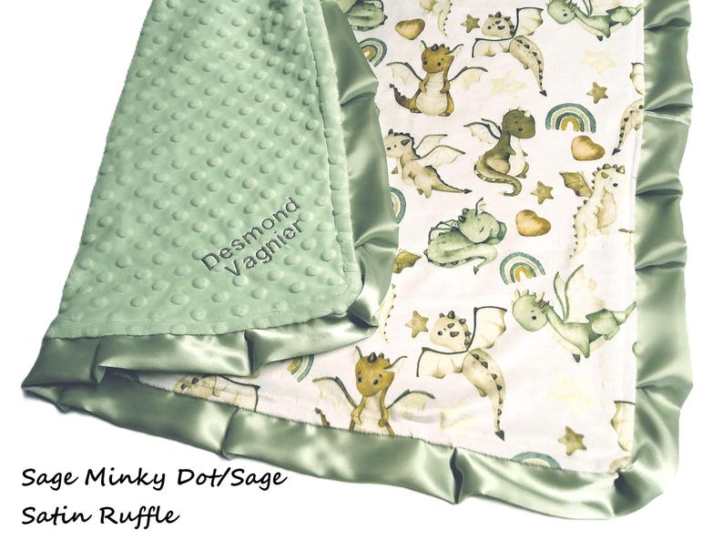May include: Sage green baby blanket with a dragon pattern. The blanket features a soft, textured minky dot fabric on one side and a dragon print on the other. The edges are trimmed with a sage satin ruffle. The name "Desmond Vagnier" is embroidered on the minky side.