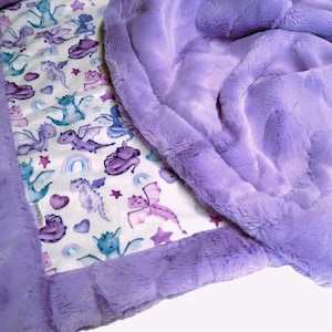 May include: A soft, purple baby blanket with a dragon pattern. The blanket features a white background with colorful dragons, rainbows, and stars. The border is a plush, textured purple material.