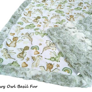 May include: A baby blanket featuring a white background with a pattern of cartoon dragons in shades of brown and green, rainbows, and stars. The blanket has a soft, green fur border. The text "Snowy Owl Basil Fur" is at the bottom.