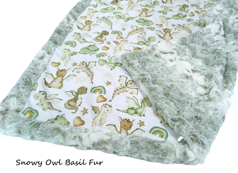 May include: A baby blanket featuring a white background with a pattern of cartoon dragons in shades of brown and green, rainbows, and stars. The blanket has a soft, green fur border. The text "Snowy Owl Basil Fur" is at the bottom.