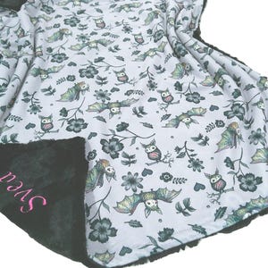 May include: A soft blanket with a white background featuring a repeating pattern of green and grey bats, owls, and flowers. The reverse side is solid black plush fabric with the name Svea embroidered in pink script lettering.