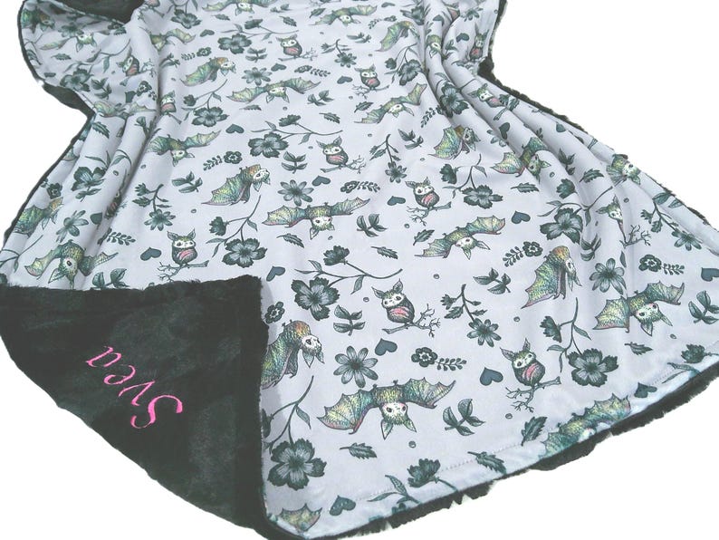 May include: A soft blanket with a white background featuring a repeating pattern of green and grey bats, owls, and flowers. The reverse side is solid black plush fabric with the name Svea embroidered in pink script lettering.