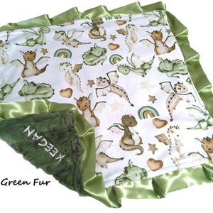 May include: A baby blanket with a white background featuring a pattern of cartoon dragons in shades of green, brown, and cream. The blanket has a green satin trim and a green fur section with the name "KEEGAN" embroidered on it. The words "Loden Green Fur" are also visible.