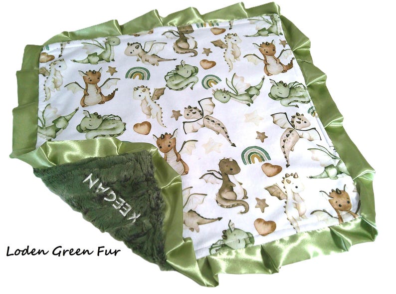 May include: A baby blanket with a white background featuring a pattern of cartoon dragons in shades of green, brown, and cream. The blanket has a green satin trim and a green fur section with the name "KEEGAN" embroidered on it. The words "Loden Green Fur" are also visible.