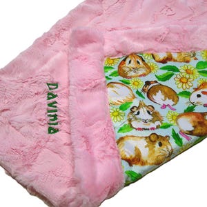 May include: A pink, plush baby blanket with the name "Davinia" embroidered in green. The blanket features a guinea pig and flower pattern on a light blue background.