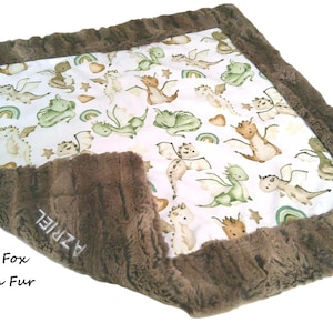 May include: A baby blanket with a white center featuring watercolor dragons, rainbows, and stars. The blanket is trimmed with a brown, textured fur. The text "AZRIEL" is embroidered on the fur. The words "Forest Fox" and "Brown Fur" are also visible.