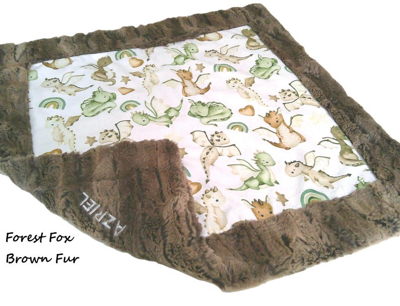 May include: A baby blanket with a white center featuring watercolor dragons, rainbows, and stars. The blanket is trimmed with a brown, textured fur. The text "AZRIEL" is embroidered on the fur. The words "Forest Fox" and "Brown Fur" are also visible.