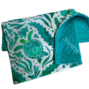May include: A baby blanket featuring a sea turtle design. The blanket has a white background with green sea plants and a turtle. The reverse side is teal with a dimpled texture. The name "Scarlett" is embroidered on the teal side.