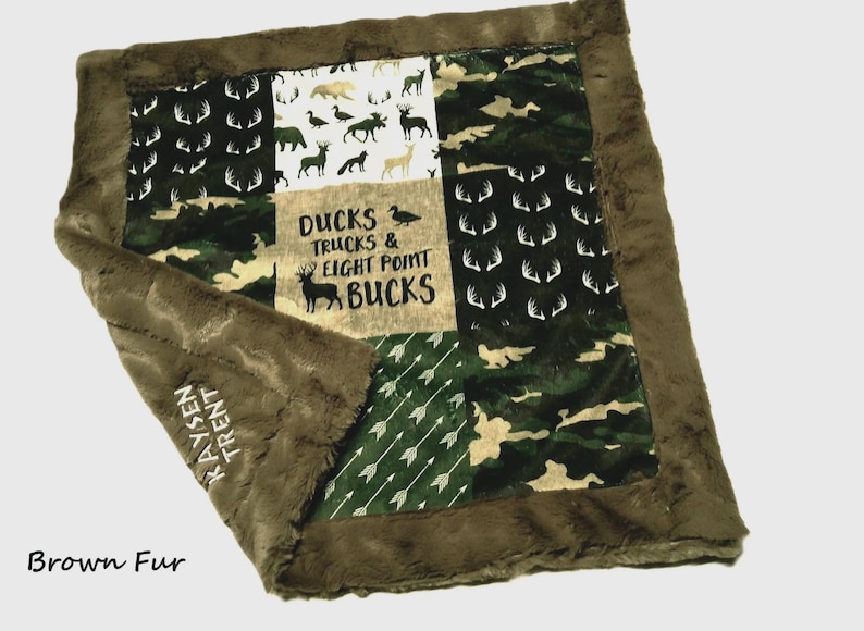 May include: A brown and green patchwork baby blanket with a soft, plush border. The blanket features various patterns, including camouflage, deer antlers, and arrows. The center panel has the text "DUCKS TRUCKS & EIGHT POINT BUCKS".