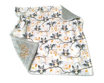 May include: A white baby blanket featuring a repeating pattern of grey bats perched on brown branches with orange autumn leaves. The blanket has a textured grey border on one corner with the name Roo embroidered in orange thread.