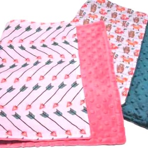 May include: Three baby blankets with different patterns. One blanket is pink with a white arrow pattern, another is white with a brown and orange animal pattern, and the third is teal.