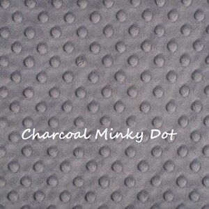May include: A charcoal gray fabric featuring a raised, embossed dot pattern across the entire surface. The text 'Charcoal Minky Dot' is printed in white in the center of the fabric, which has rounded corners and a soft, textured appearance.