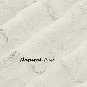 May include: Close-up of a cream-colored faux fur fabric with a soft, textured appearance. The fabric has a wavy pattern and the words "Natural Fur" are printed on the surface.
