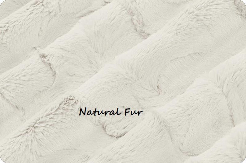 May include: Close-up of a cream-colored faux fur fabric with a soft, textured appearance. The fabric has a wavy pattern and the words "Natural Fur" are printed on the surface.