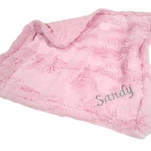 May include: A soft, light pink plush blanket featuring a textured, swirling faux fur surface on one side and a dimpled minky fabric on the reverse. The blanket is personalized with the name Sandy embroidered in a grey script font in the corner.