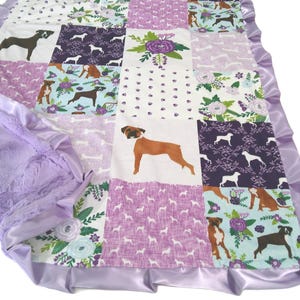 May include: A patchwork baby blanket featuring various dog breeds in brown and black on a white, purple, and teal background. The blanket has a soft, textured purple backing and a satin lavender trim.