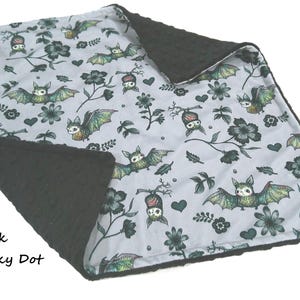 May include: A double-sided blanket featuring a light gray top layer with a repeating pattern of green and black bats, flowers, and hearts. The reverse side is a solid black minky dot fabric with a raised, textured circular pattern throughout.