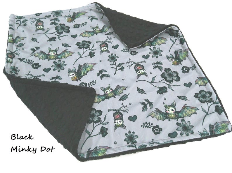 May include: A double-sided blanket featuring a light gray top layer with a repeating pattern of green and black bats, flowers, and hearts. The reverse side is a solid black minky dot fabric with a raised, textured circular pattern throughout.