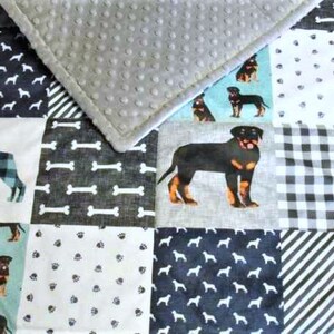 May include: A patchwork dog-themed blanket featuring various patterns, including dog silhouettes, bones, paw prints, and a large Rottweiler image. The blanket has a gray, textured border and a soft, gray, dotted backing.