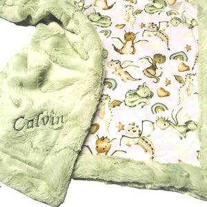 May include: A soft, light green baby blanket with a plush, textured surface. The blanket features an embroidered name, "Calvin," and a patterned interior with cartoon dragons, rainbows, and stars. The edges are trimmed with a fluffy border.