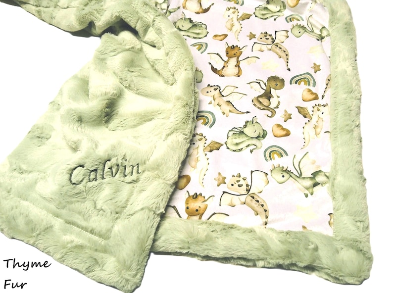 May include: A soft, light green baby blanket with a plush, textured surface. The blanket features an embroidered name, "Calvin," and a patterned interior with cartoon dragons, rainbows, and stars. The edges are trimmed with a fluffy border.