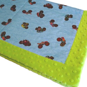 May include: A baby blanket with a light blue background featuring a pattern of monster trucks in various colors. The blanket has a bright green, textured border.