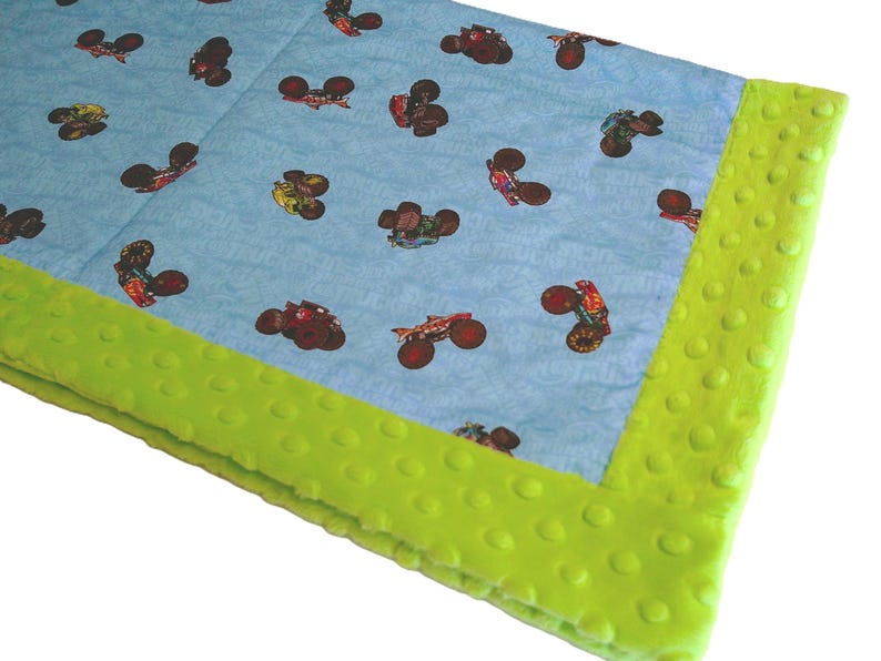 May include: A baby blanket with a light blue background featuring a pattern of monster trucks in various colors. The blanket has a bright green, textured border.