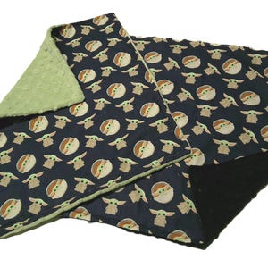 May include: A navy blue baby blanket featuring a repeating pattern of a cartoon character. The blanket has a green, textured backing. The edges are trimmed with green fabric. The blanket is folded.