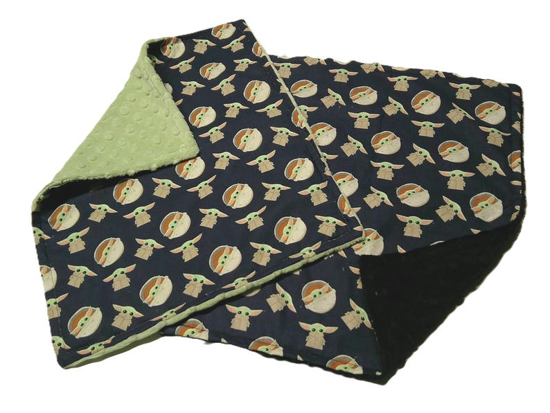 May include: A navy blue baby blanket featuring a repeating pattern of a cartoon character. The blanket has a green, textured backing. The edges are trimmed with green fabric. The blanket is folded.