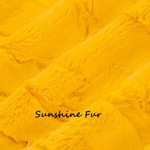 Pu&ograve; includere: Primo piano di un tessuto in finta pelliccia giallo brillante. Il materiale morbido presenta un motivo strutturato e ondulato. Le parole "Sunshine Fur" sono stampate in nero, in minuscolo.