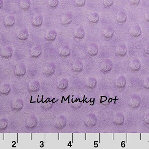 May include: A lilac-colored minky fabric featuring a raised dot texture across the surface. The text "Lilac Minky Dot" is printed in the center. A white ruler at the bottom shows measurements in inches, spanning from 1 to 8 inches in length.