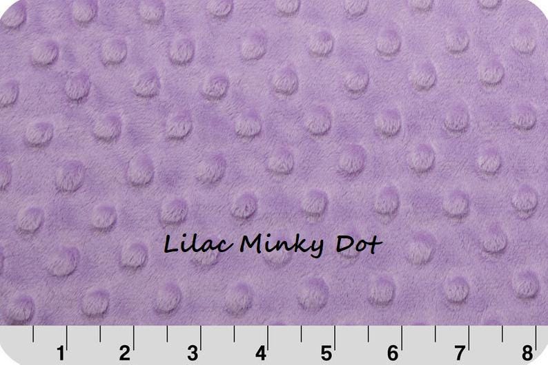 May include: A lilac-colored minky fabric featuring a raised dot texture across the surface. The text "Lilac Minky Dot" is printed in the center. A white ruler at the bottom shows measurements in inches, spanning from 1 to 8 inches in length.