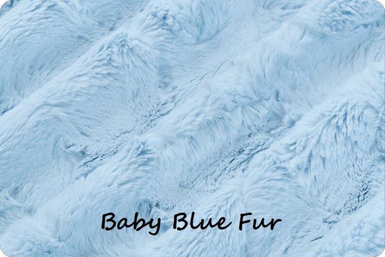 May include: A soft, light blue faux fur fabric with a dense, plush texture and wavy, directional pile. The surface features a consistent baby blue color throughout, with the text "Baby Blue Fur" printed in black in the lower center area.